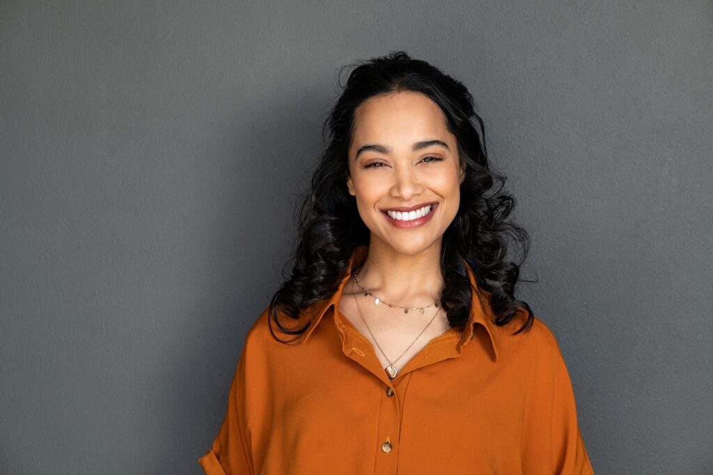 Confident Woman Smiling - Ultra Dental A cheerful woman with curly hair in a burnt orange blouse smiling confidently against a gray background - Ultra Dental.