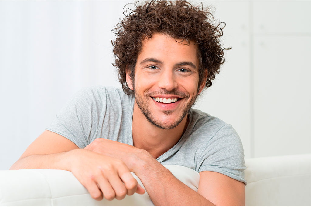 Young Man with Healthy Smile - Ultra Dental A cheerful young man with curly hair smiling while leaning on a couch - Ultra Dental.