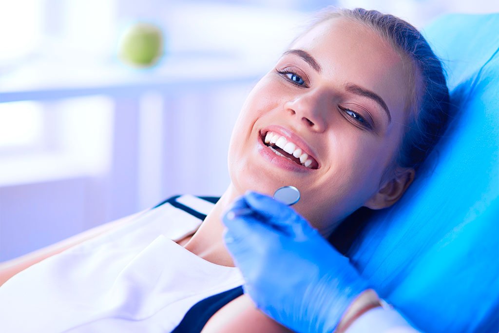 Relaxed Woman at Dental Appointment - Ultra Dental A young woman reclining in a dental chair smiling while being examined with a dental mirror - Ultra Dental.