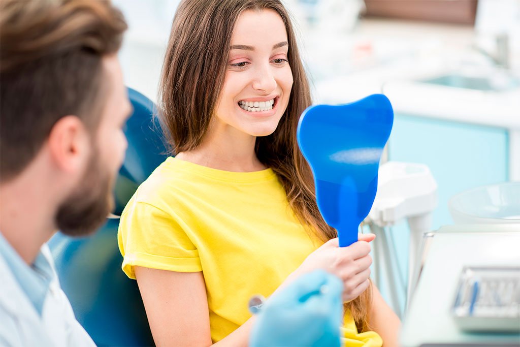 Patient Checking Smile in Mirror A female dental patient smiling into a blue tooth-shaped mirror during her appointment