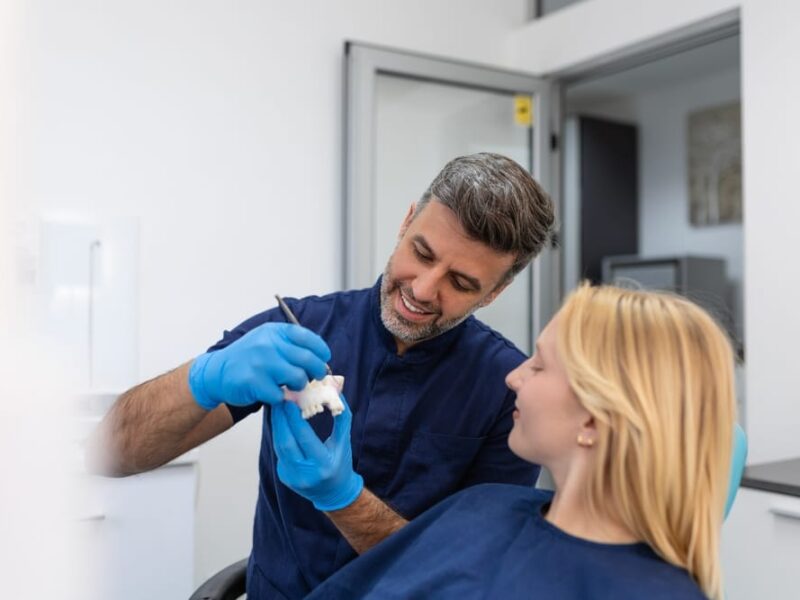 Dentist explaining a dental bridge procedure to a female patient using a dental model in a modern clinic setting.