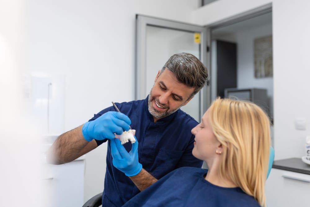 Dentist explaining a dental bridge procedure to a female patient using a dental model in a modern clinic setting.