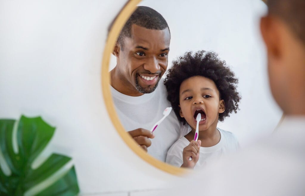 A dad and child brush their teeth together, showing the importance of building healthy dental habits early.
