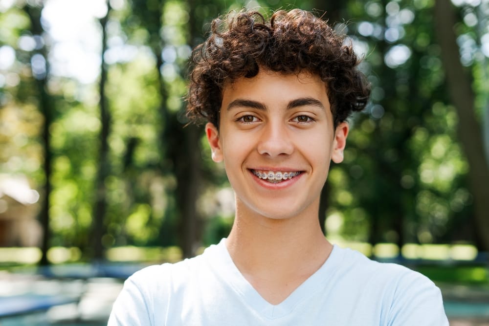 Smiling teenage boy with curly hair wearing braces, standing outdoors in a park setting.