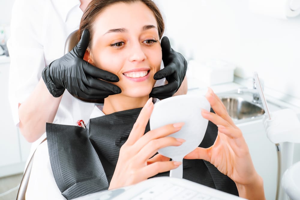 A woman sits in the dental chair holding a mirror while a dental professional adjusts her smile, highlighting her satisfaction with the results – Dental Implants in San Fernando, CA.