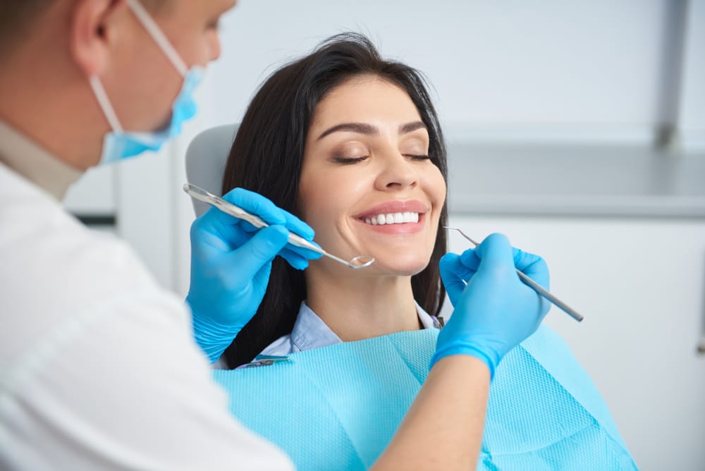 A woman sits at home holding her cheek with visible discomfort, indicating possible tooth pain or a dental emergency requiring urgent care – Emergency Dentist in San Fernando, CA.