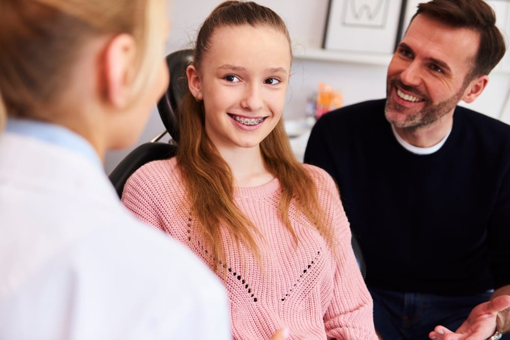 Teen Girl with Braces Talking to Dentist with Parent Beside Her – Dentist in Pacoima, CA A teenage girl with braces smiles while speaking with a dental professional, accompanied by her supportive parent, highlighting a positive orthodontic visit – Dentist in Pacoima, CA.