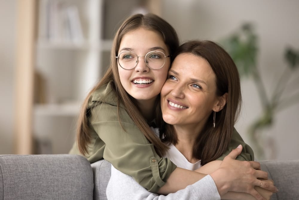 A cheerful woman with curly hair and glasses relaxes at home, enjoying a warm drink while smiling brightly, showcasing confidence and comfort – Orthodontist Sylmar.