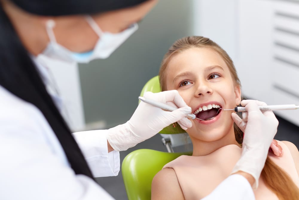 A smiling young girl sits in the dental chair while a pediatric dentist gently cleans her teeth, creating a positive and reassuring dental experience – Pediatric Dentist in San Fernando.