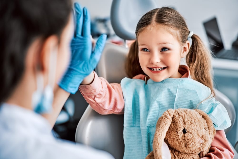 A cheerful young girl with pigtails gives her dentist a high-five while holding a stuffed bunny, celebrating a successful and fun dental appointment – Pediatric Dentist in San Fernando.