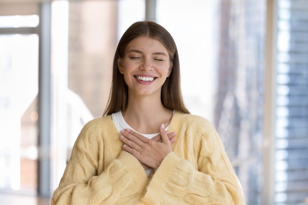 Young Woman Holding Hands to Chest, Smiling with Gratitude – Dentist in Pacoima, CA A serene young woman embraces a moment of gratitude with her hands placed on her chest, eyes closed, and a bright smile that reflects happiness and wellness – Dentist in Pacoima, CA.
