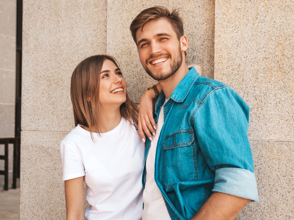 Happy Couple Standing Together Outdoors – Dentist in Pacoima, CA A cheerful couple stands close together, smiling brightly while leaning against a light stone wall, capturing a moment of connection and confidence – Dentist in Pacoima, CA.